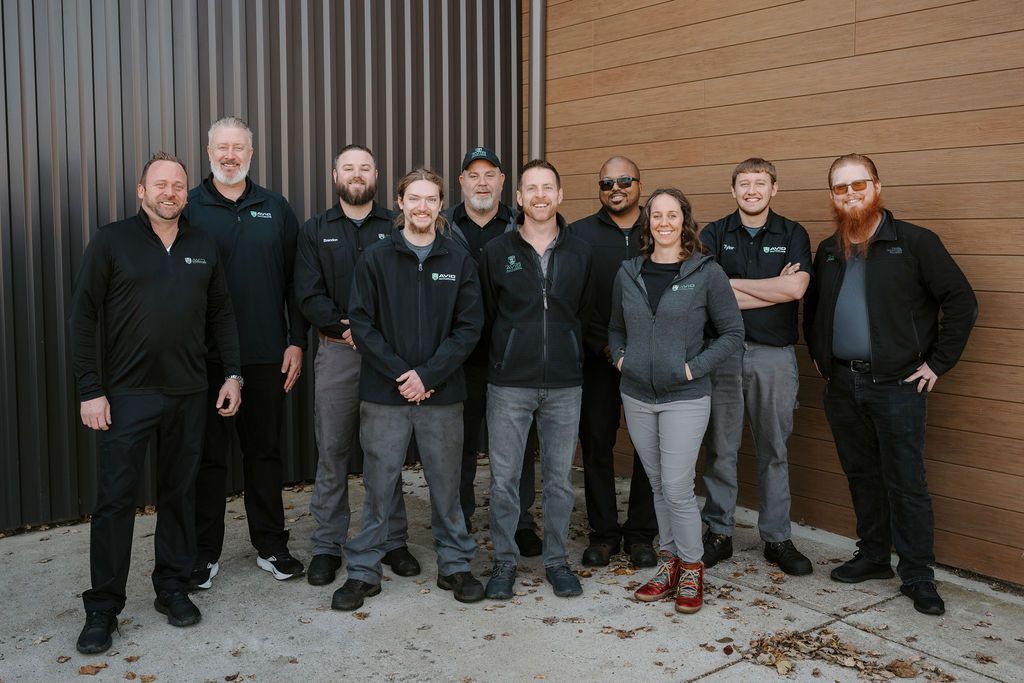 Nine people stand together in front of a wooden wall, wearing mostly dark jackets and casual clothes.