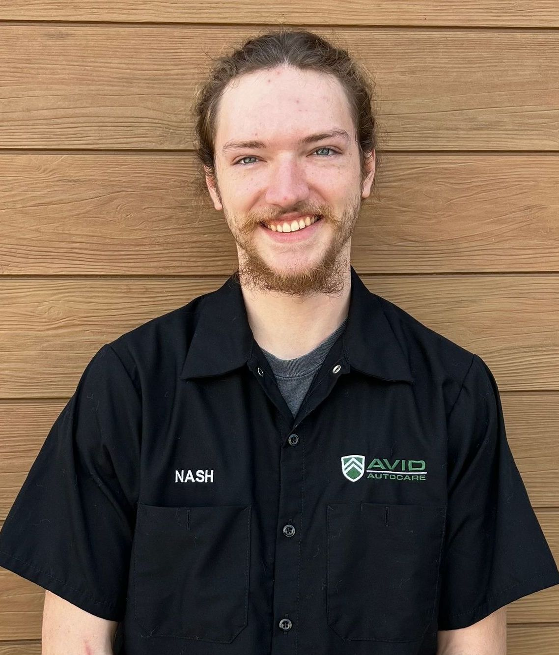 Smiling person in a black work shirt standing against a wooden wall