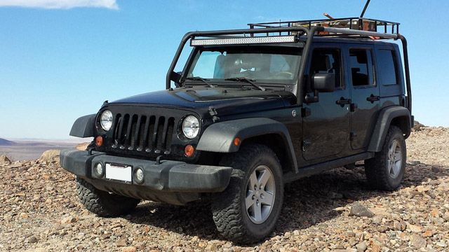 Black Jeep Wrangler parked on rocky terrain under a clear sky, with roof rack and off-road gear.