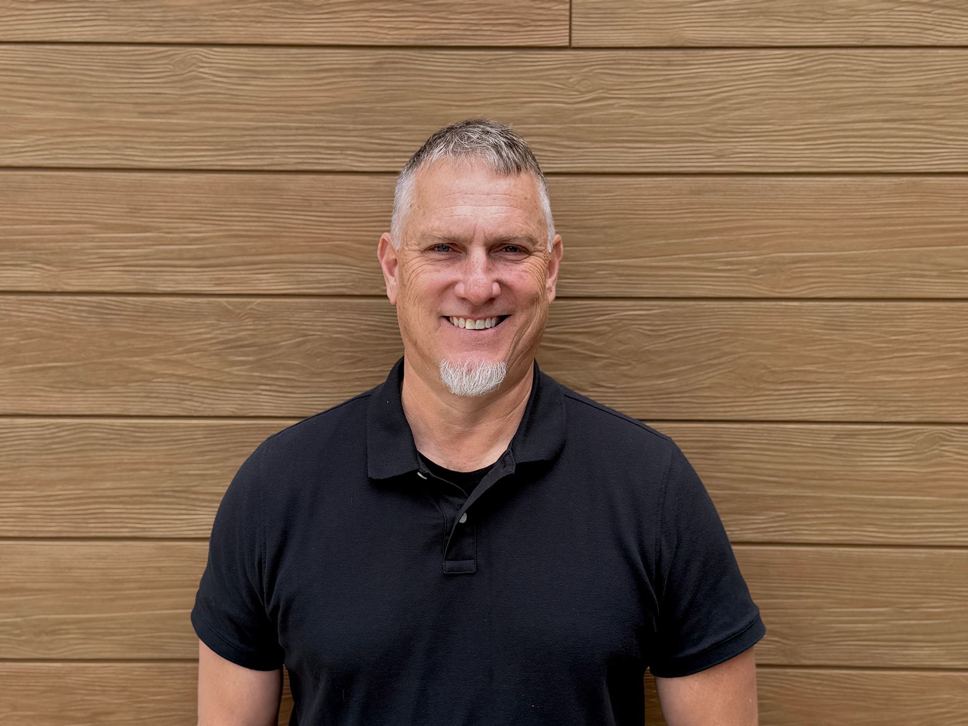 Smiling man in a black polo shirt standing in front of a wooden wall