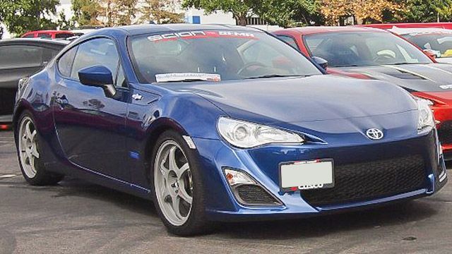 Blue sports coupe parked outdoors in a lot, front-left view, with other cars in the background