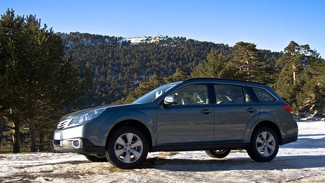 Silver station wagon parked on a snowy road with pine trees and mountains in the background