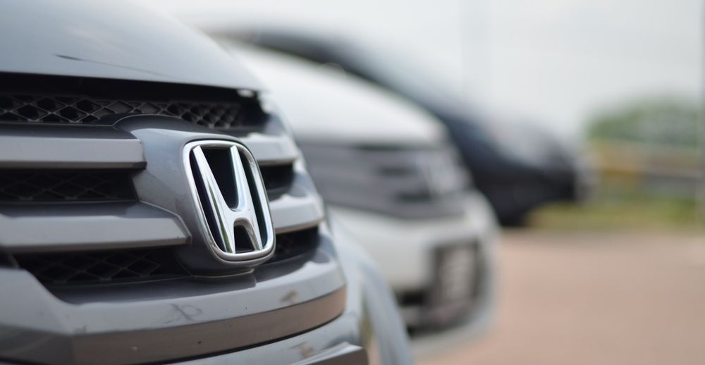 Close-up of a silver Honda car grille with a blurred row of parked vehicles in the background