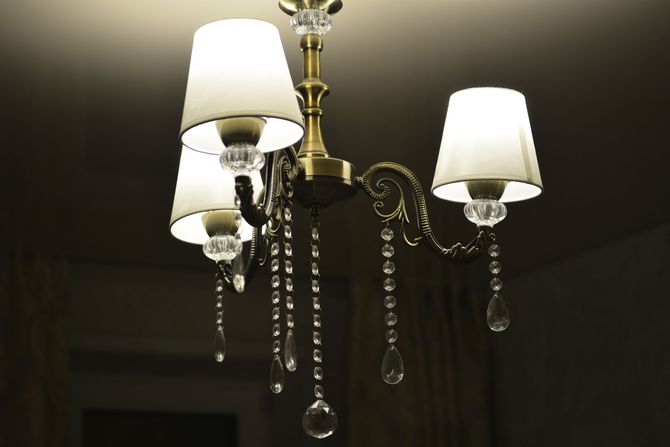 Crystal chandelier with four cream lampshades and dangling glass prisms lit against a dark ceiling.