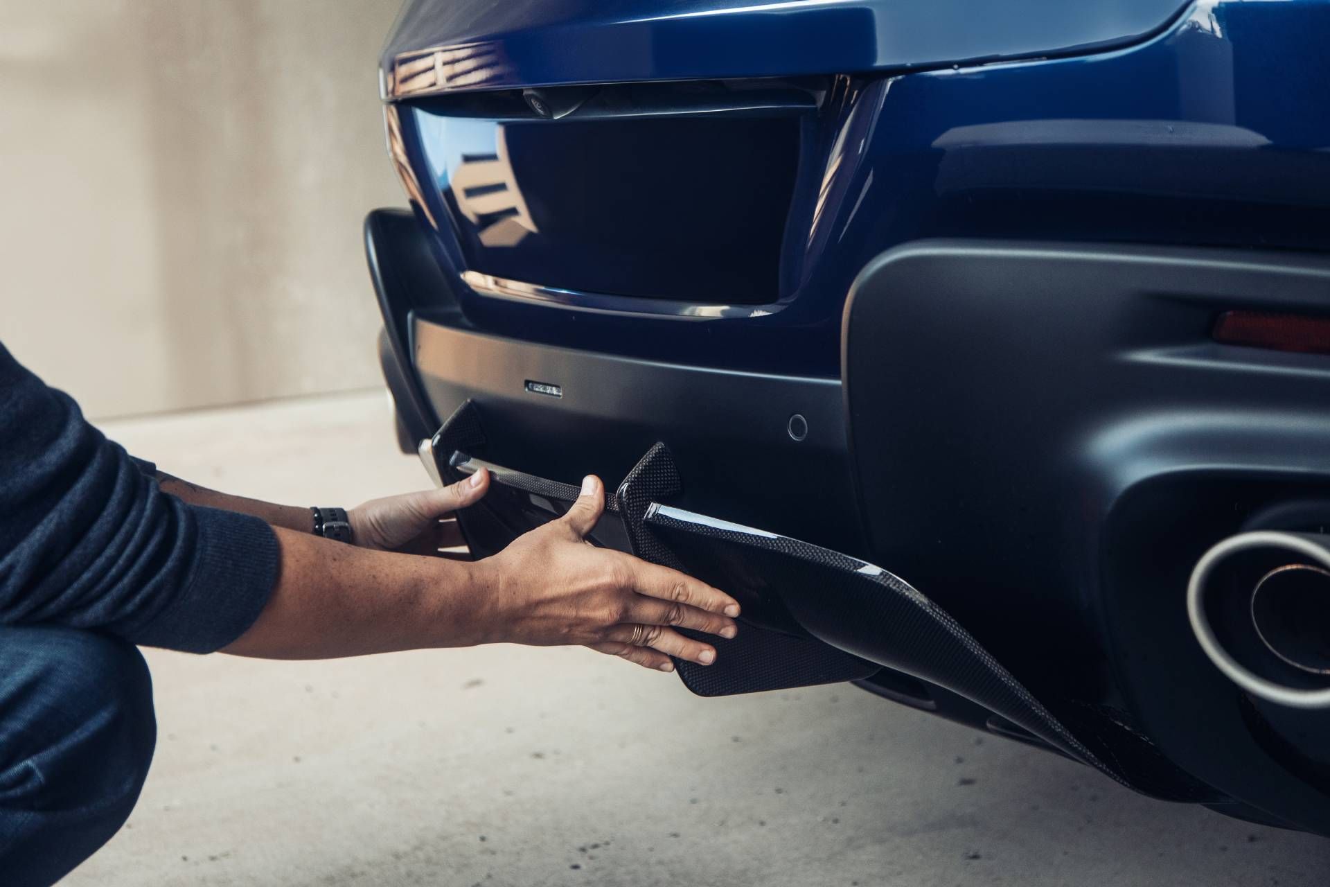 Hands under a blue car’s rear bumper, adjusting the lower trim or diffuser near the exhaust.