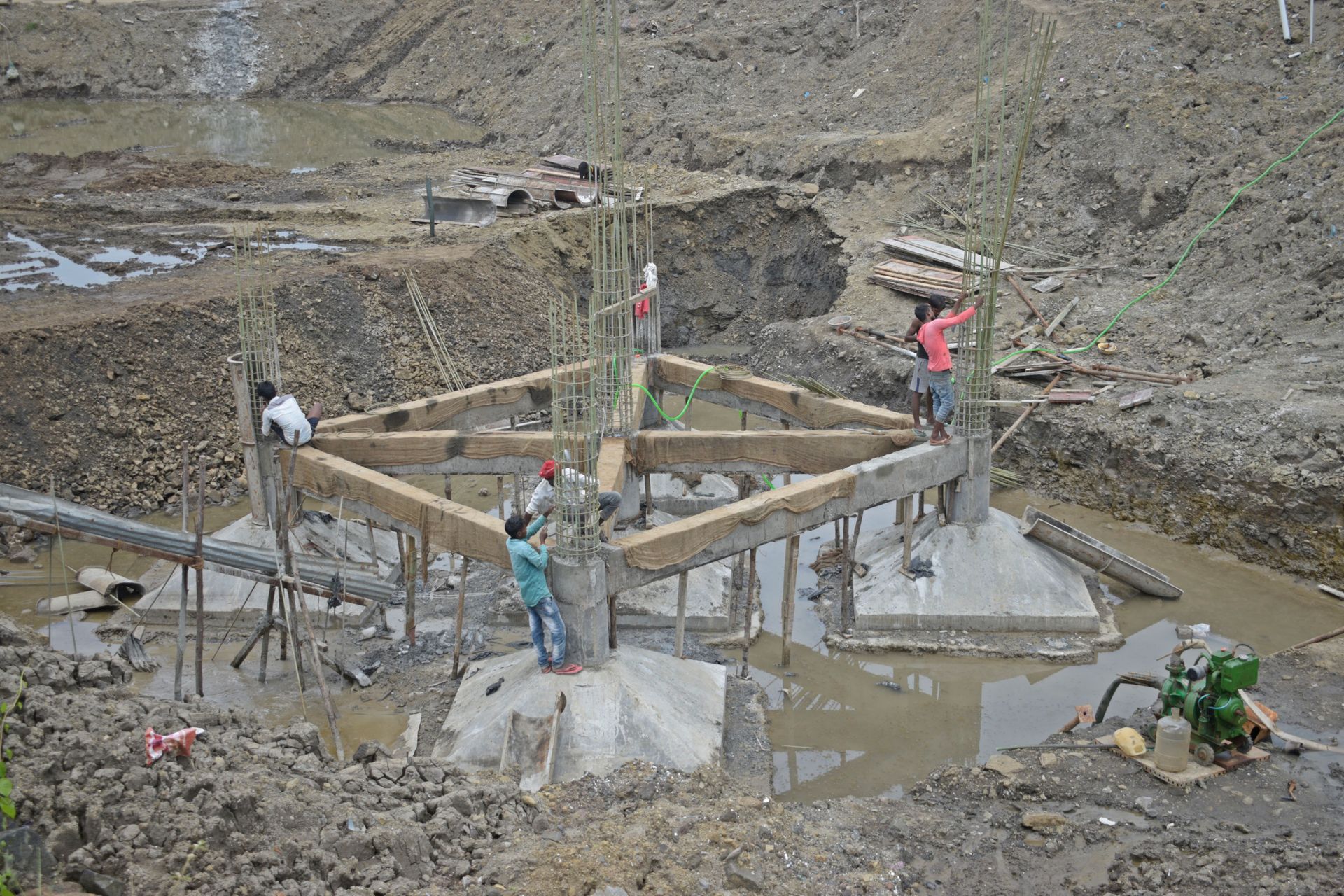 Workers build a concrete foundation in a muddy construction pit, with wooden supports and water nearby.