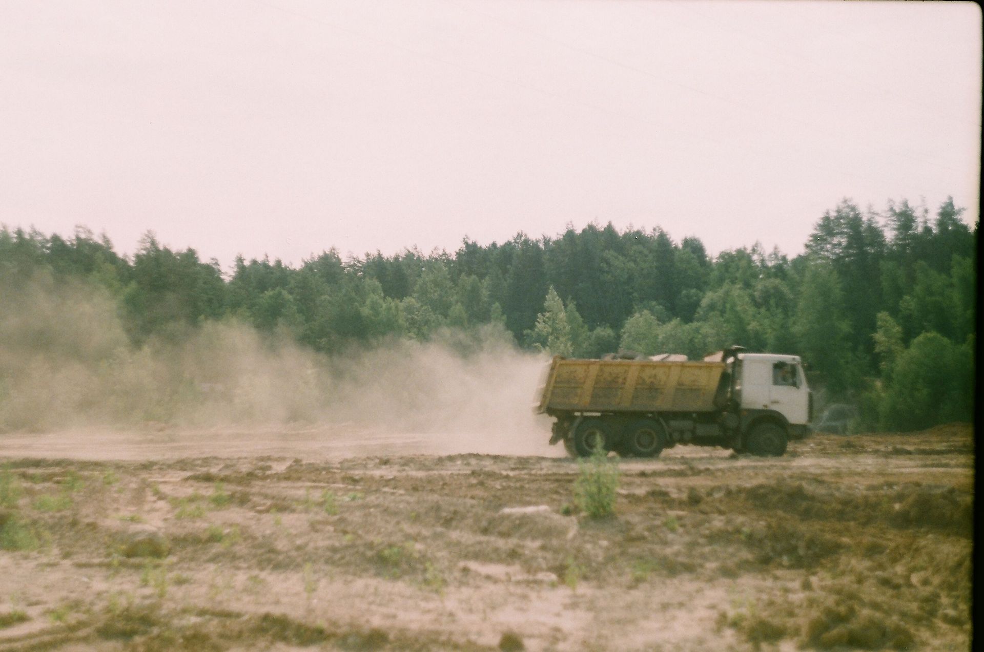 Yellow dump truck kicking up dust on a dirt road beside a forest.