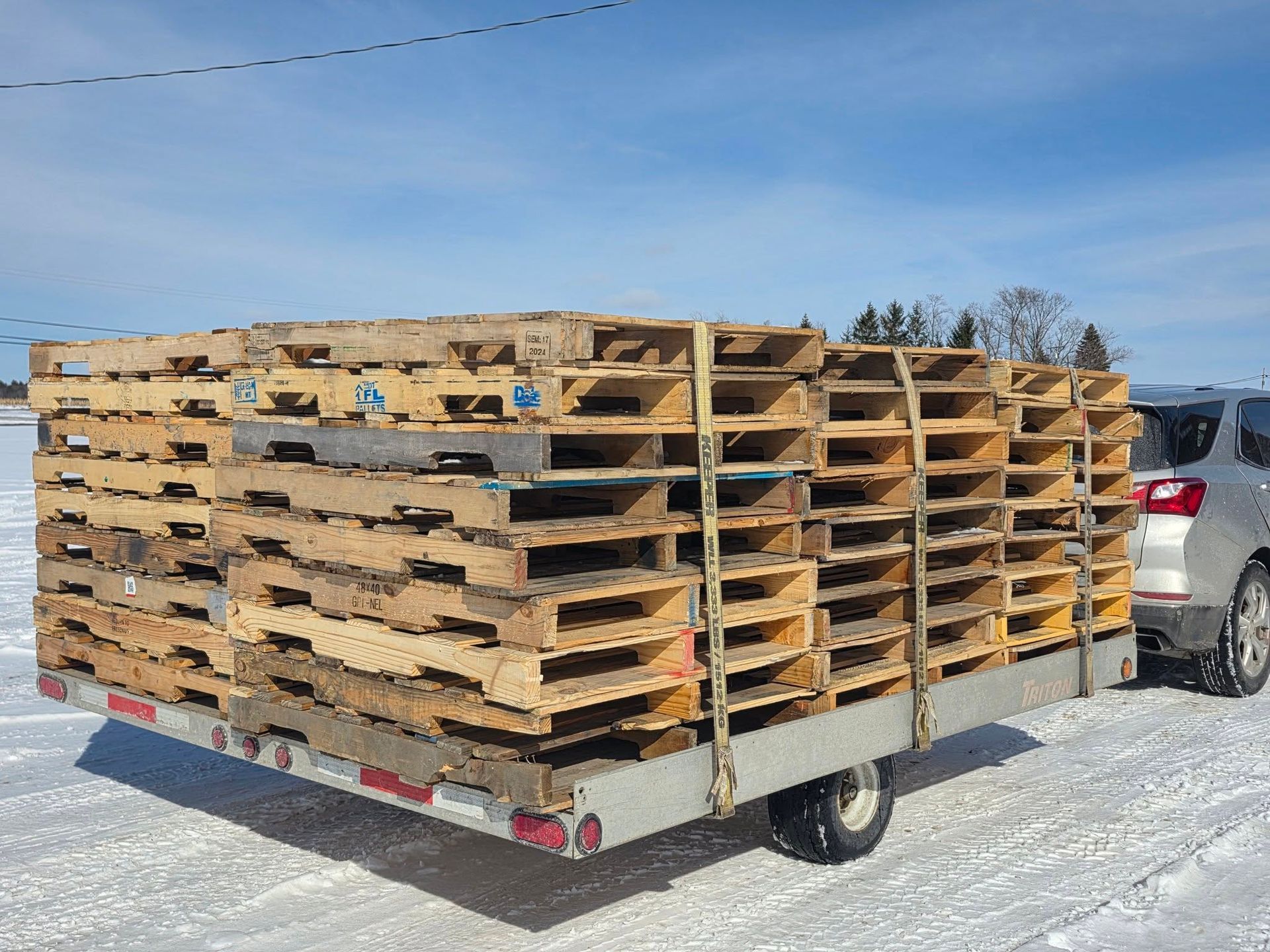 Stacked wooden pallets on a flatbed trailer in a snowy outdoor lot, with a car beside it.