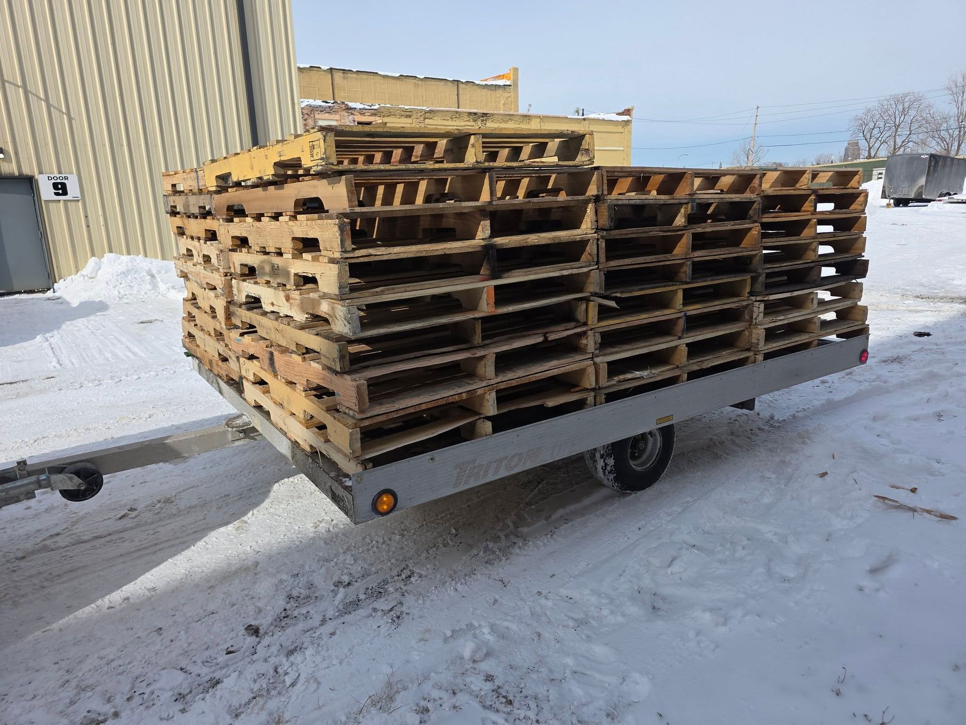 Stacked wooden pallets on a flatbed trailer in a snowy industrial lot.