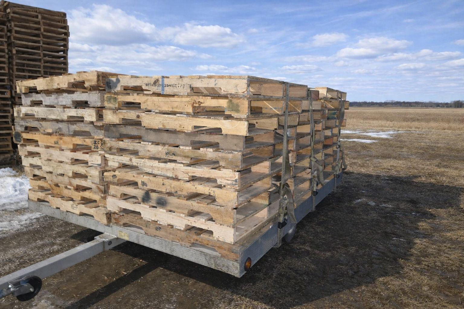 Stacked wooden pallets on a trailer in an open field, with muddy ground and a distant horizon.