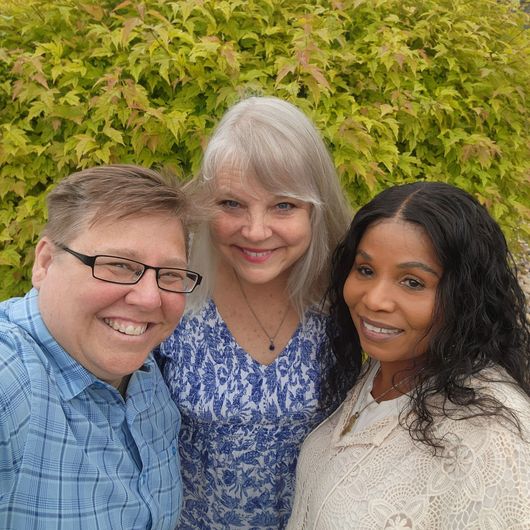 Three smiling people posing outdoors in front of green foliage, one in blue, one in white, one in blue shirt