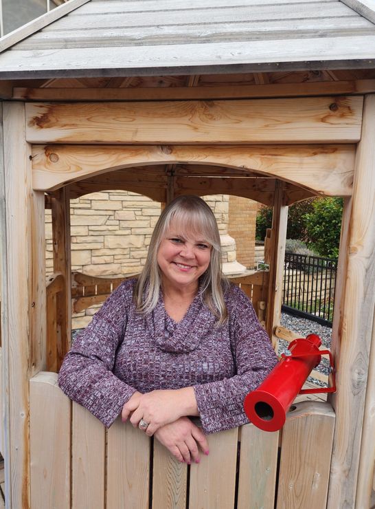 A smiling person in a purple patterned top leans on a wooden structure featuring a red toy telescope.