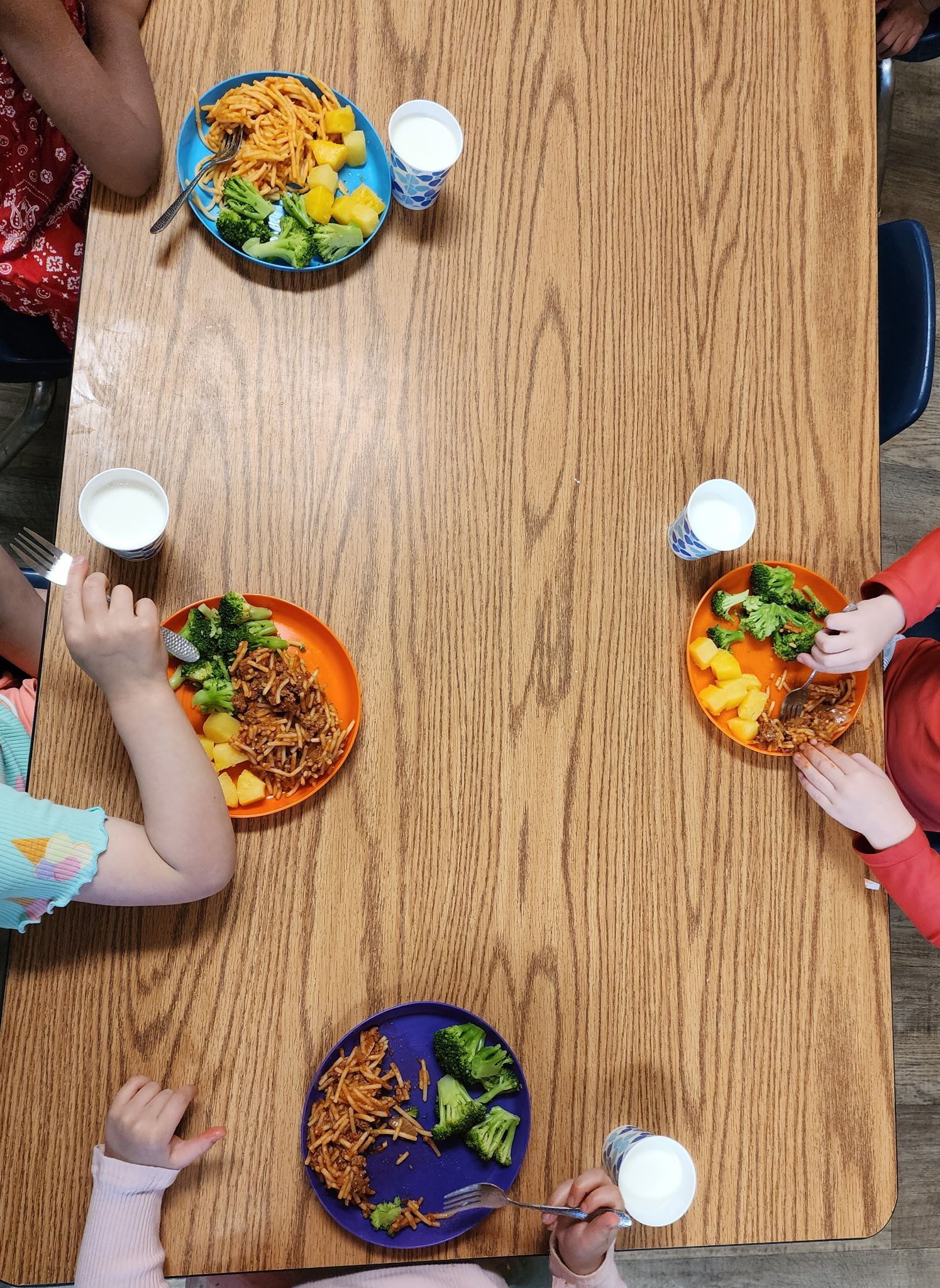 Children eating colorful meals around a wooden table in a top-down classroom setting