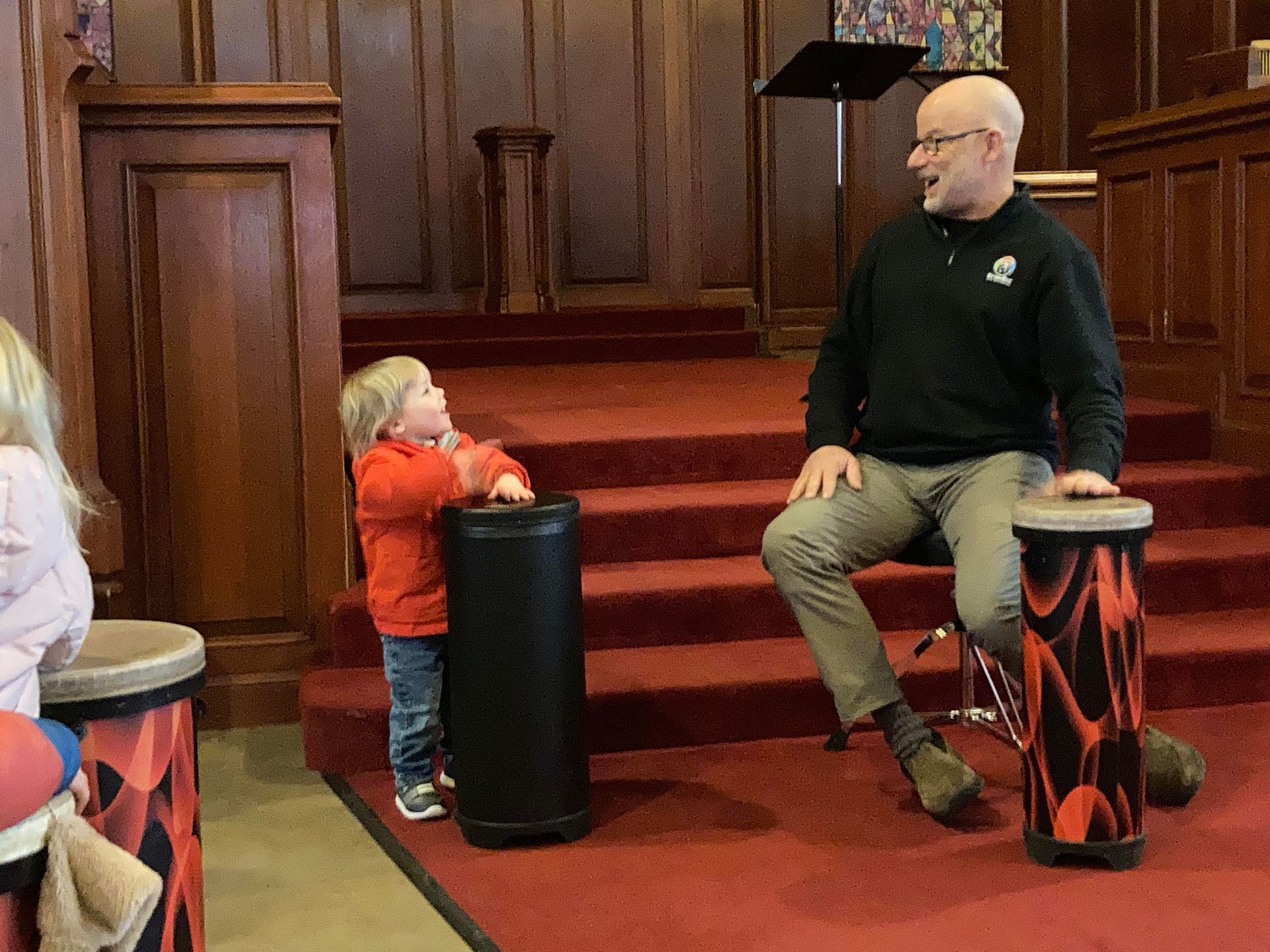 Man and child play drums on a stage with a red carpet. The man is seated, the child stands, looking up.