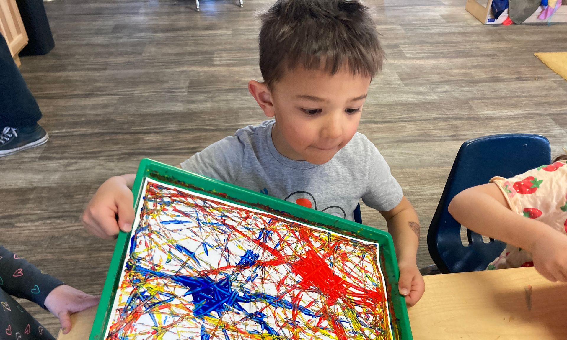 A child holds a green tray with a piece of paper featuring colorful abstract paint splatters in a classroom setting.