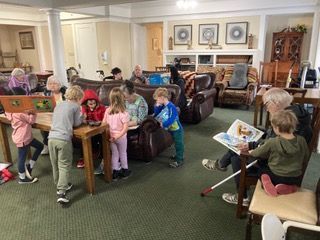 Children and seniors in a community room. Kids gather around a table and with a senior reading a book.