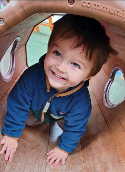 Smiling toddler on a small wooden slide, wearing blue shirt, jeans, and gray shoes. Indoor setting.