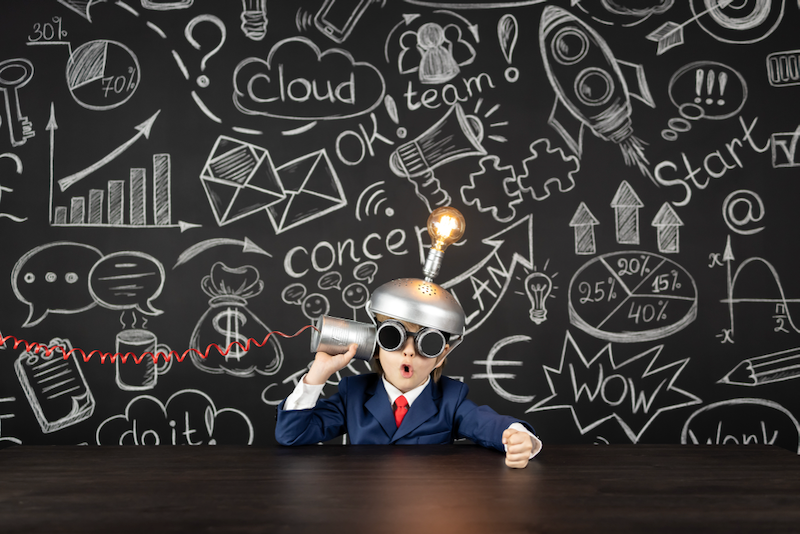 A little boy in a suit and tie is sitting at a table holding a megaphone in front of a blackboard.