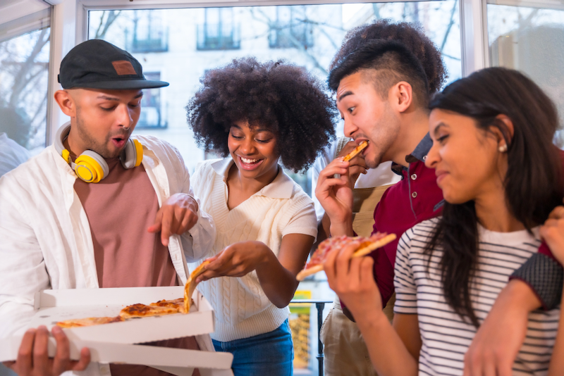 Four people eating pizza, gathered by a window. One holds the pizza box, others are eating, smiling.