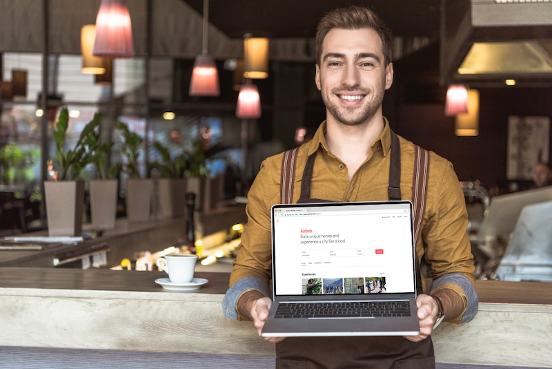 A man in an apron is holding a laptop in a restaurant.