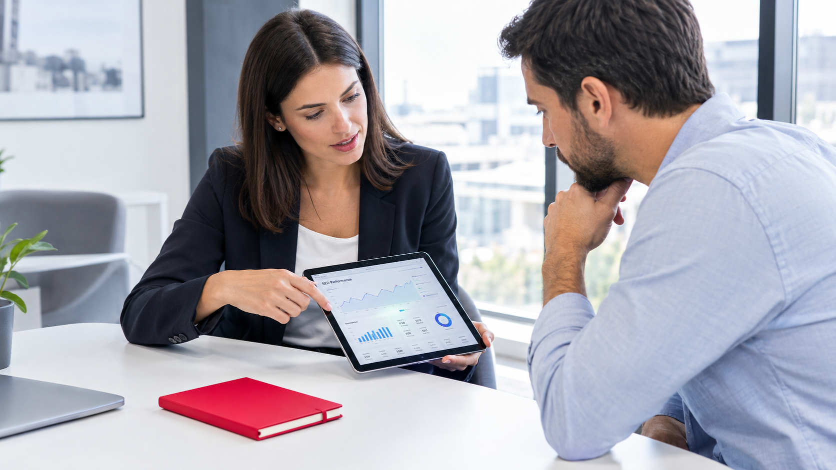 Two coworkers review a tablet at a desk, with a red notebook in the foreground.