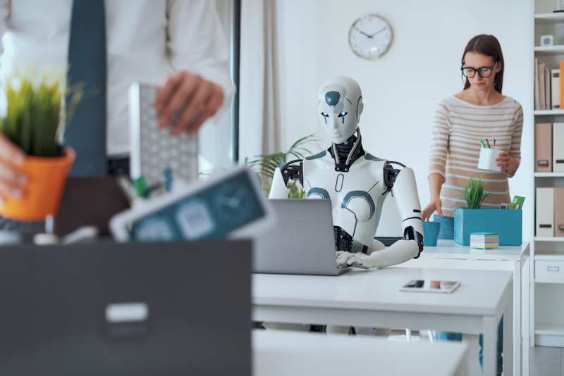A robot is sitting at a desk in front of a laptop computer.