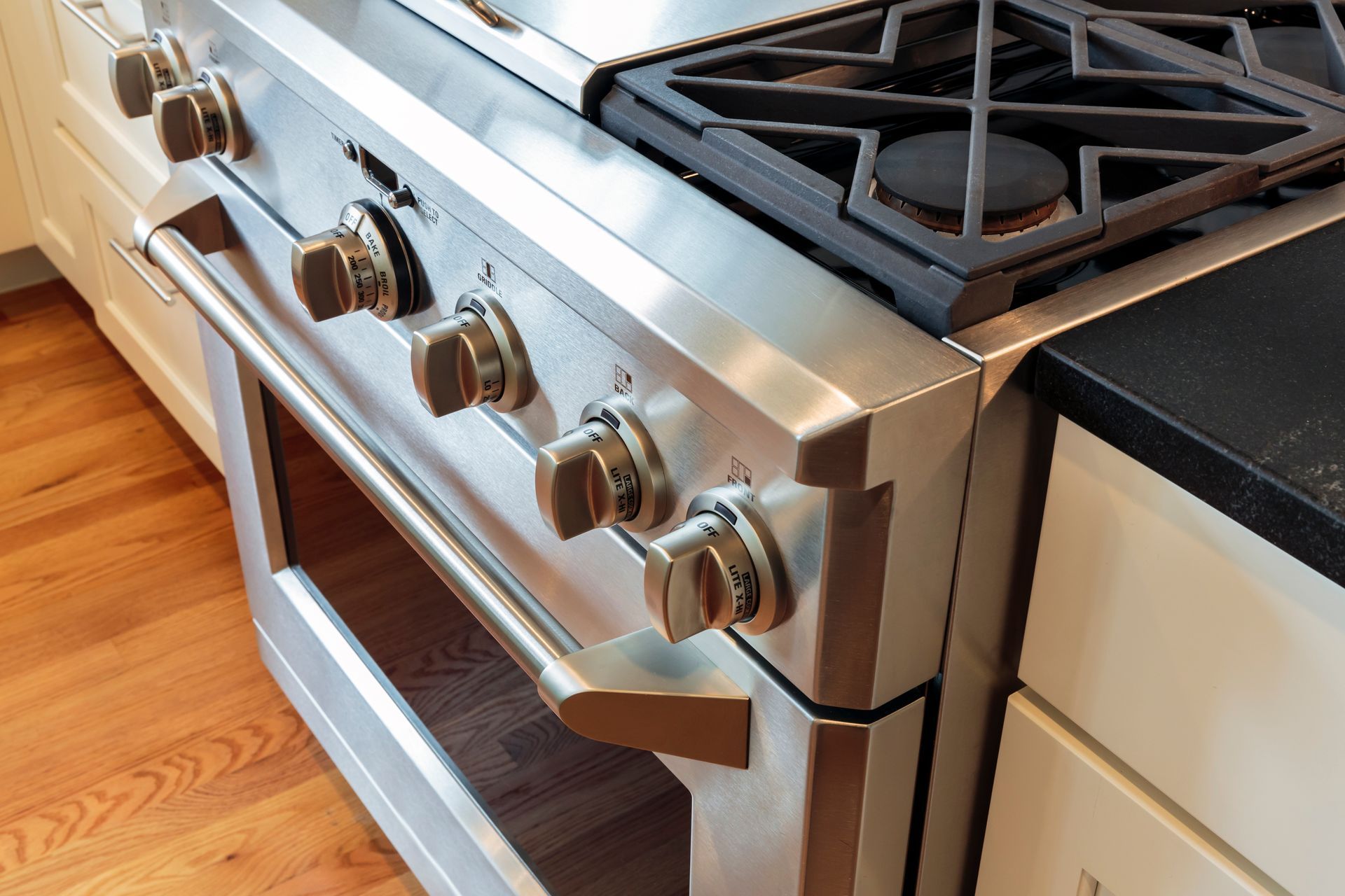 Stainless steel gas range with five knobs and black grates beside a white cabinet on a hardwood floor