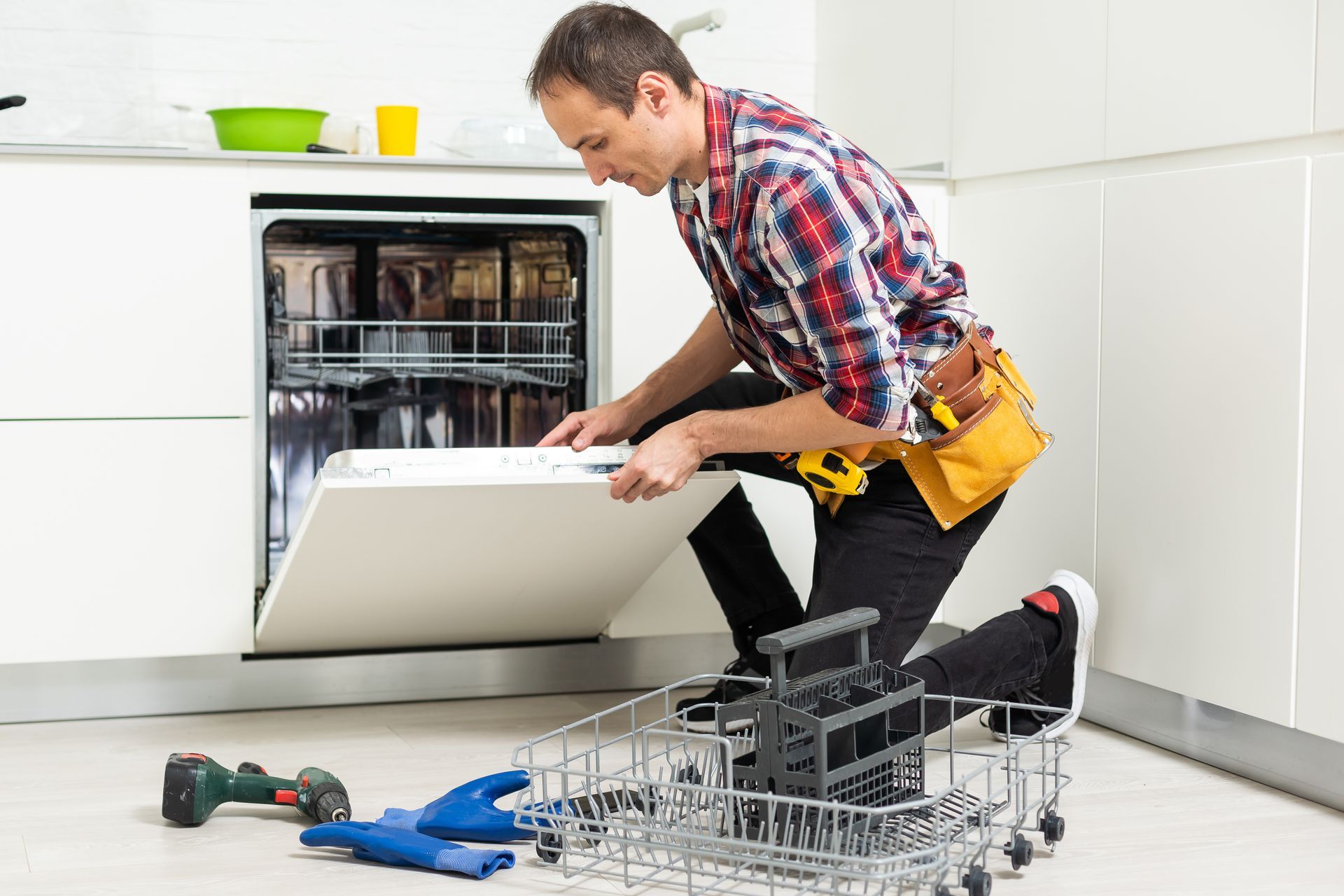 Technician repairing an open dishwasher in a kitchen, with tools and disassembled racks nearby
