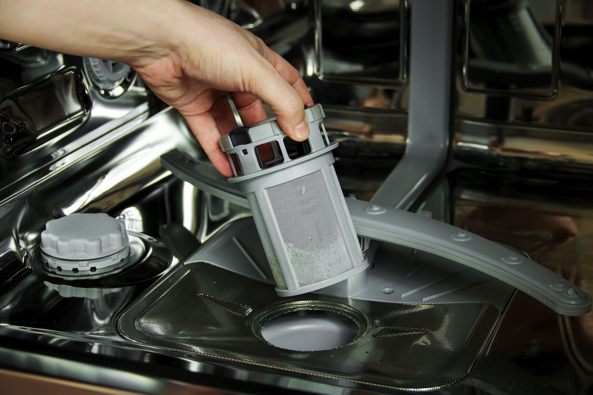 Hand placing a dishwasher filter basket back into the bottom rack of a stainless steel dishwasher
