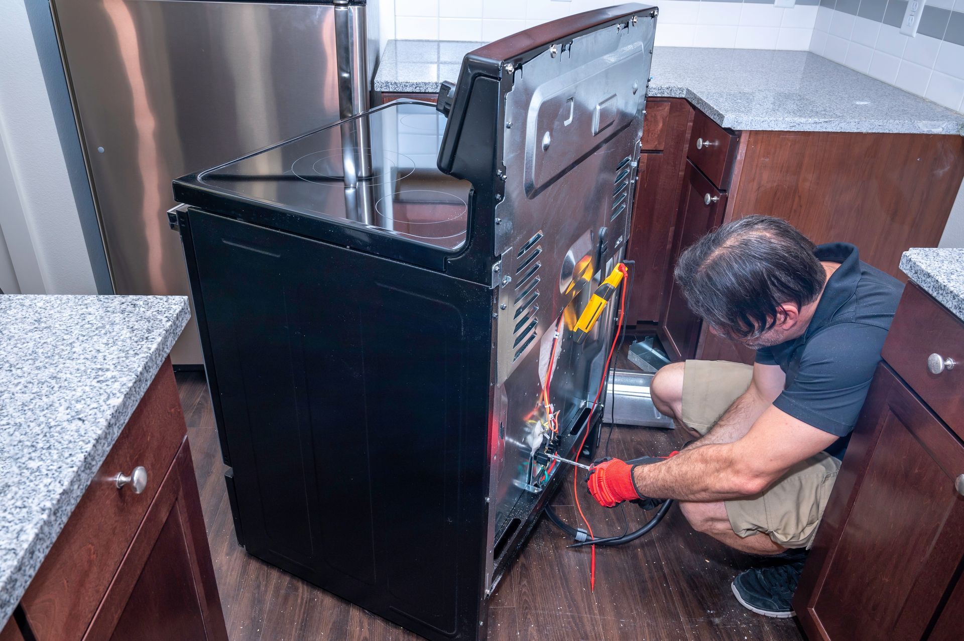 Person repairing a black oven in a kitchen, crouched beside it with tools and wiring exposed