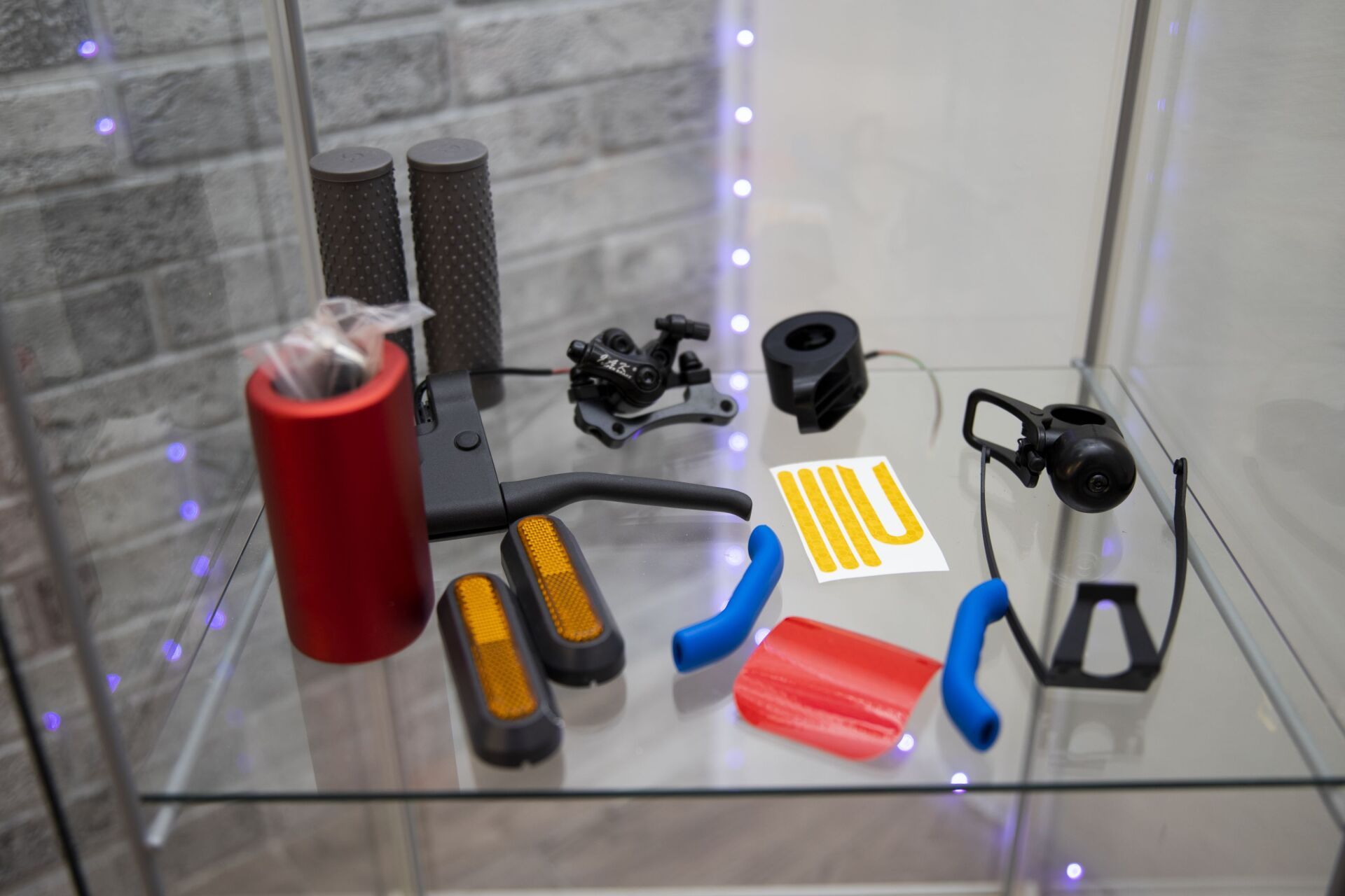 Diversas piezas y accesorios para patinetes eléctricos, como puños, reflectores y una palanca de freno, dispuestos en una estantería de cristal.