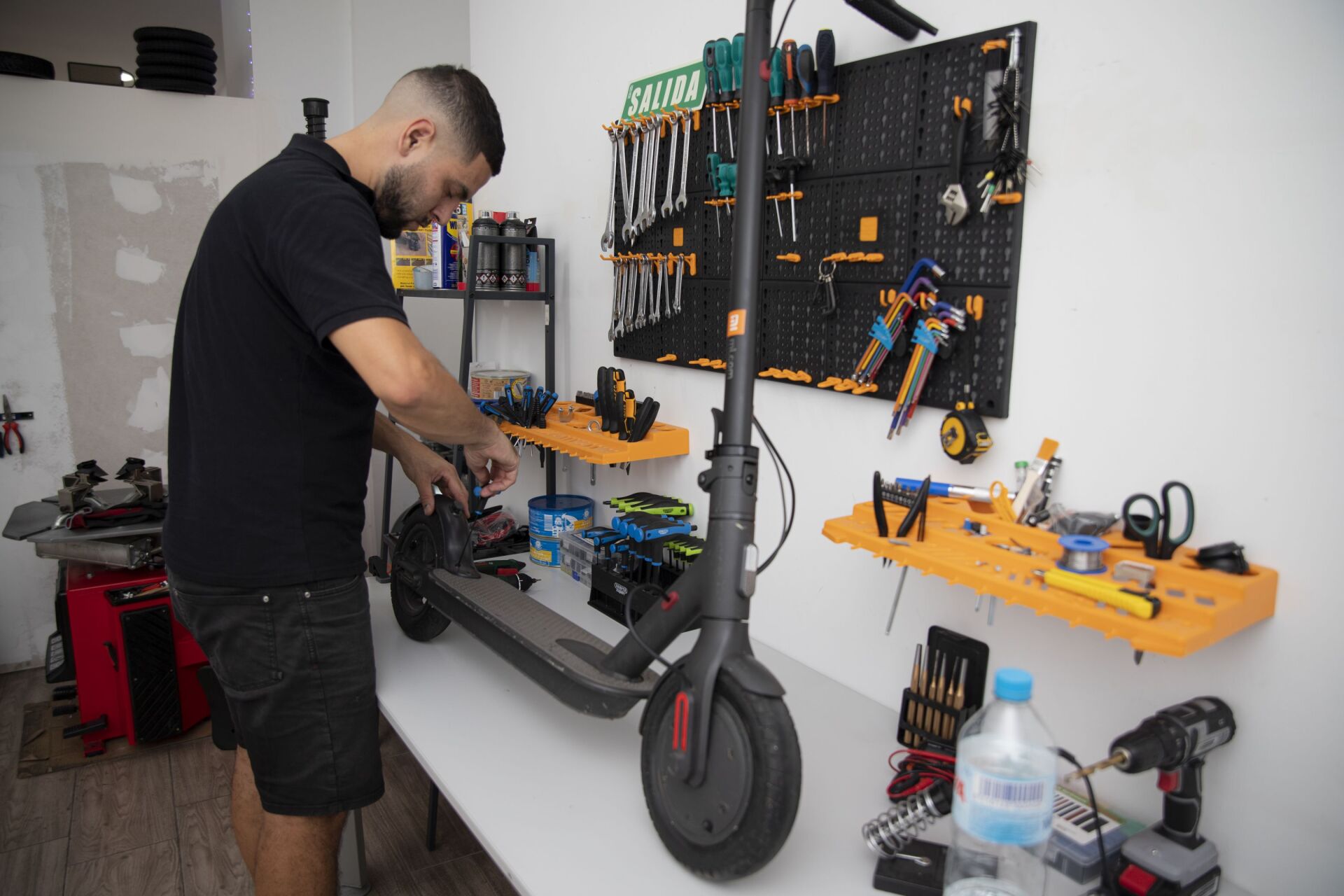 Un técnico repara un patinete eléctrico en un banco de trabajo en un taller repleto de herramientas colocadas en paneles perforados montados en la pared.