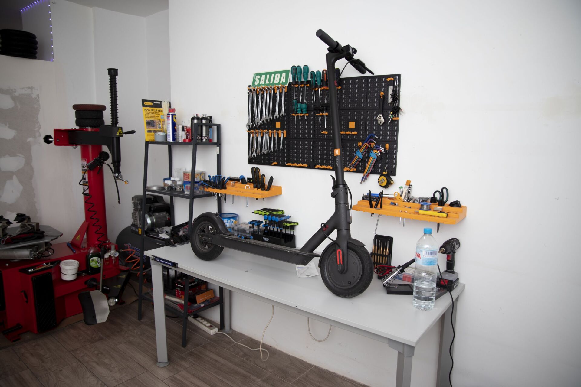 En un taller, un patinete eléctrico oscuro reposa sobre un banco de trabajo, con un panel de herramientas montado en la pared y una máquina roja para cambiar neumáticos cerca.