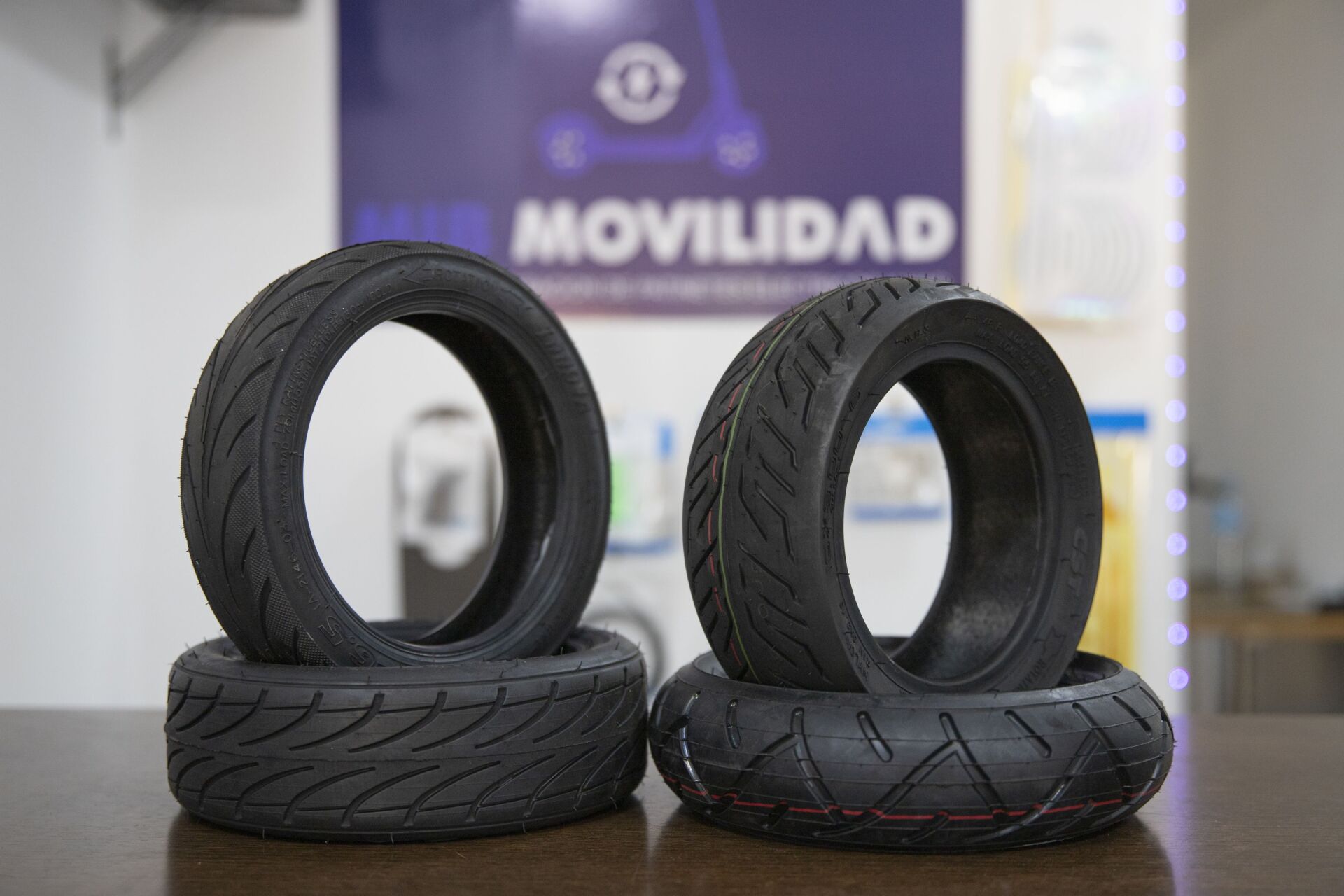 Cuatro neumáticos negros de patinete eléctrico apilados de dos en dos sobre una mesa, con un letrero de tienda borroso al fondo.