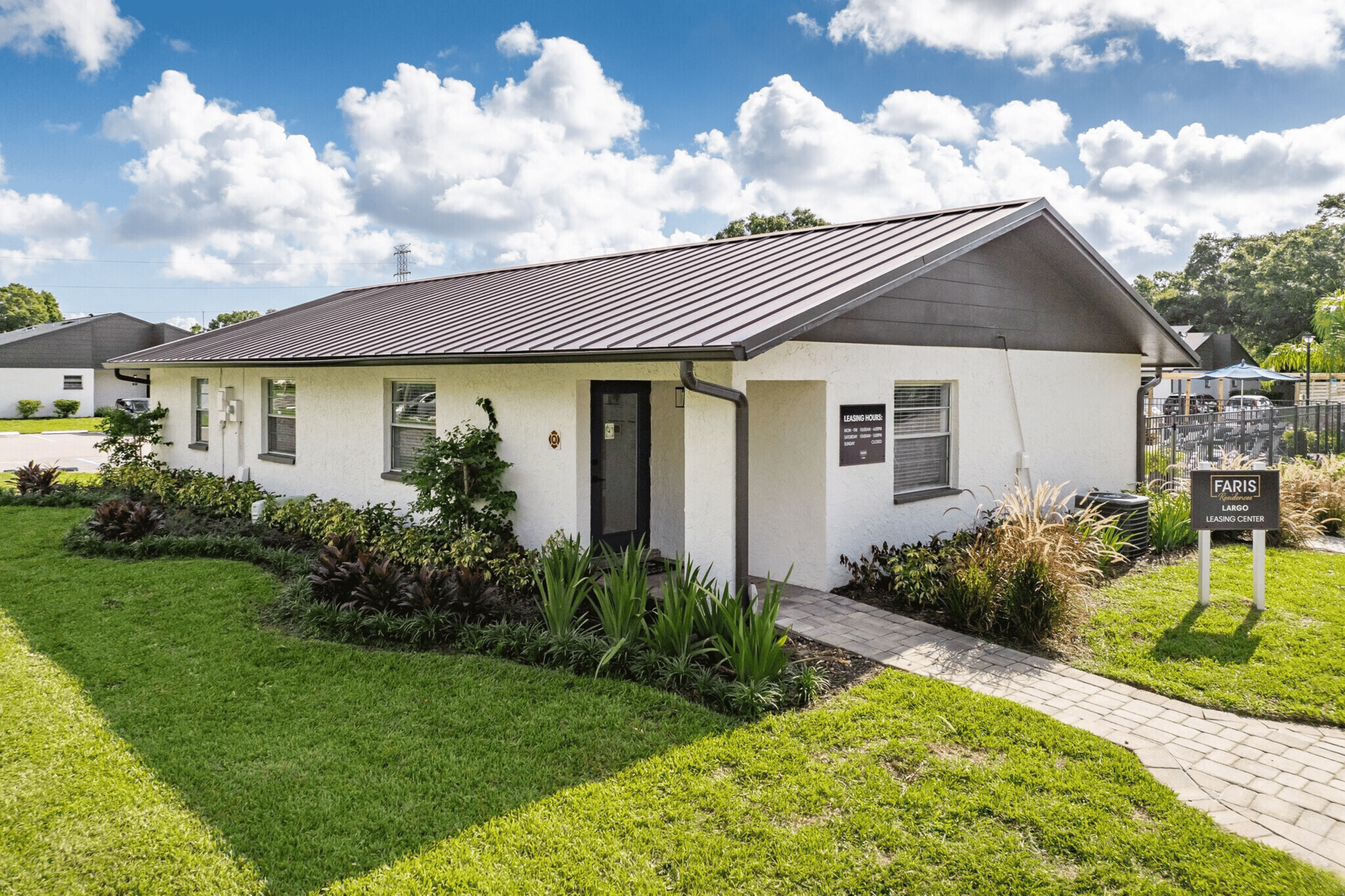 White single-story house with a tiled roof, green lawn, and a paved walkway under a partly cloudy sky