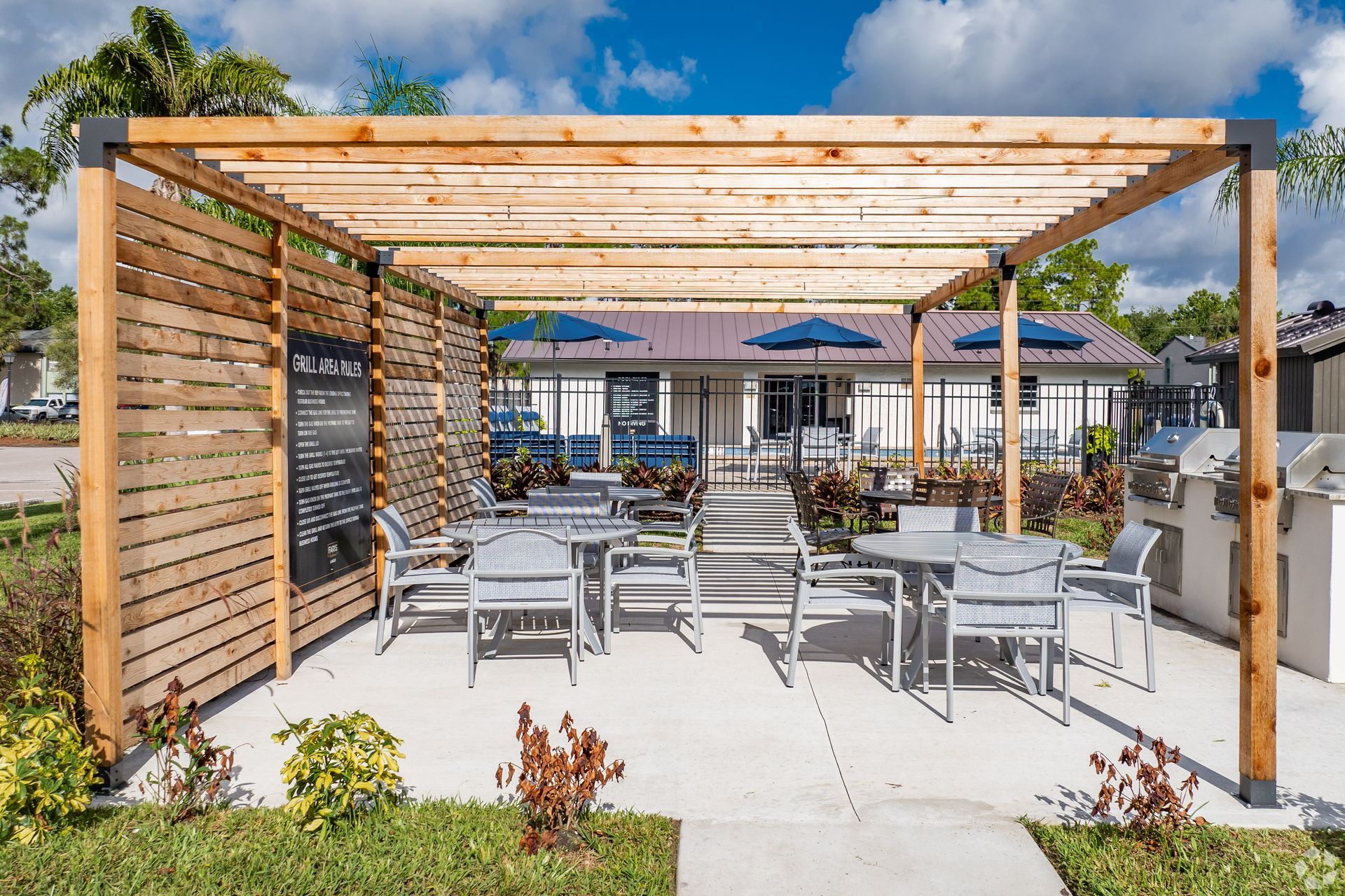 Outdoor patio seating under a wooden pergola with white tables and chairs, beside a sunny pool area