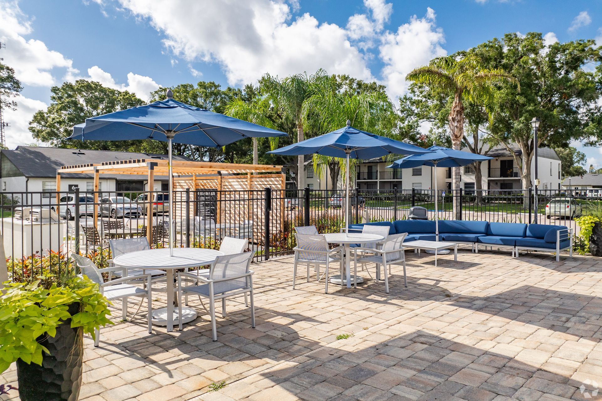 Outdoor patio with white tables, blue umbrellas, and lounge seating beside a pool under a sunny sky