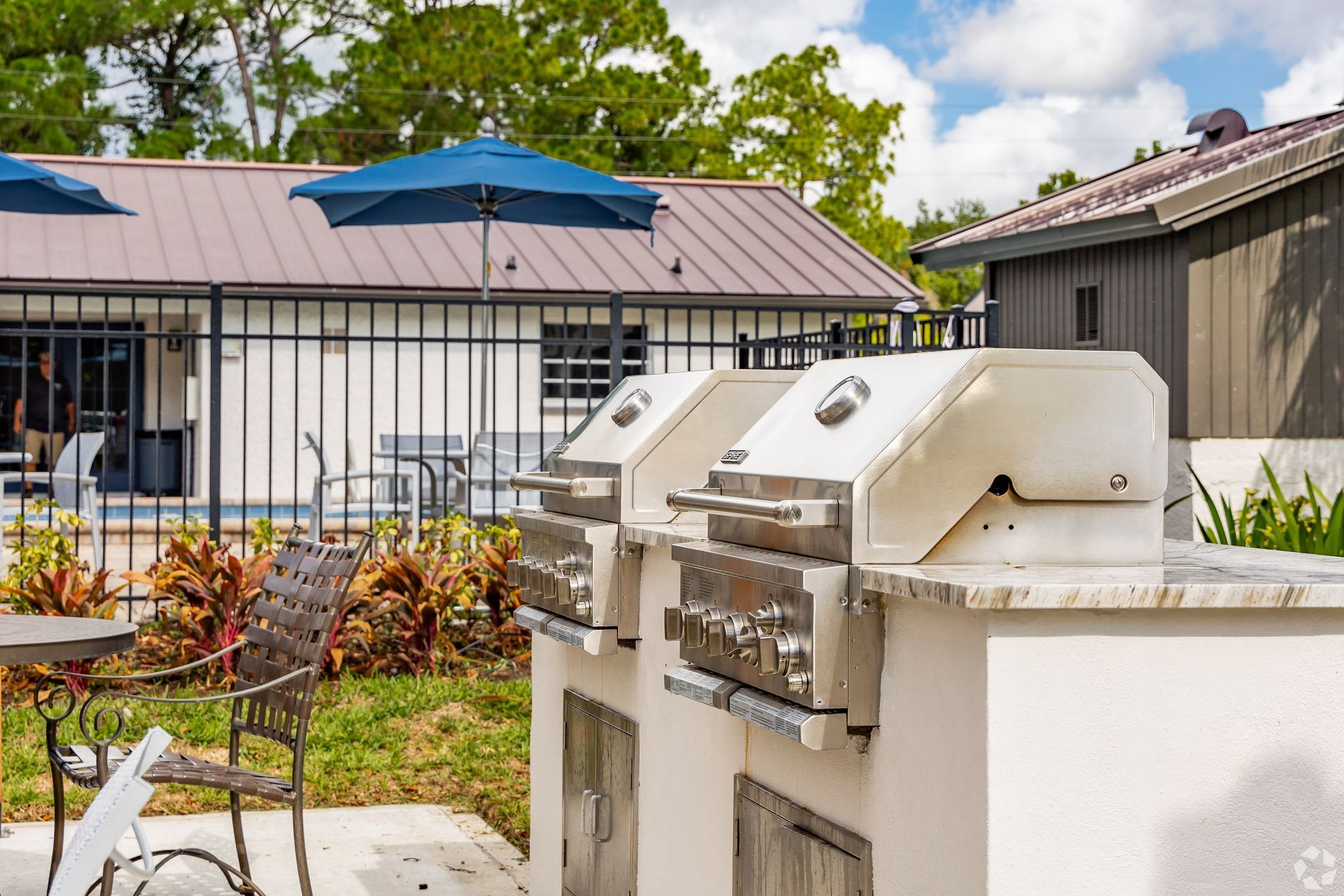 Outdoor grill and patio furniture in a fenced backyard with a house and blue umbrellas.