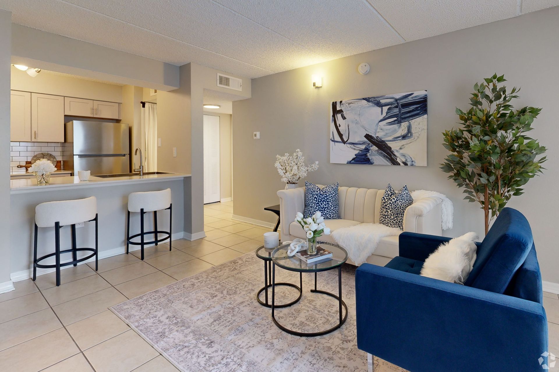 Bright modern living room with blue sofa, white armchair, coffee tables, and open kitchen bar stools
