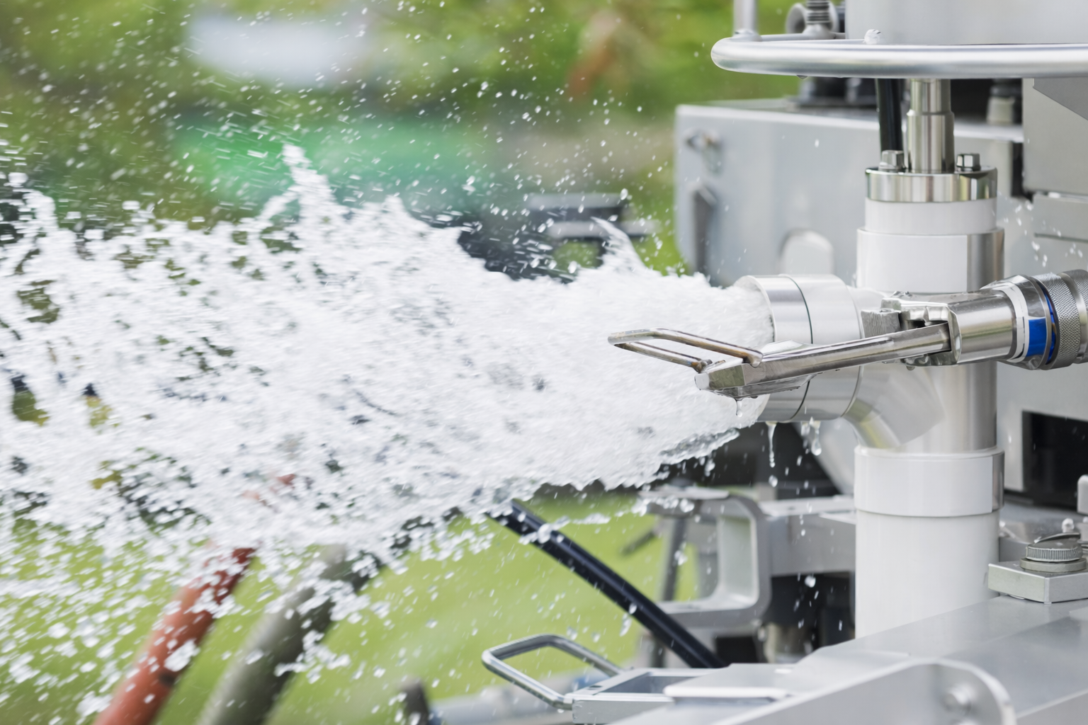 A metal industrial nozzle sprays a powerful stream of white water against a blurry green outdoor background.