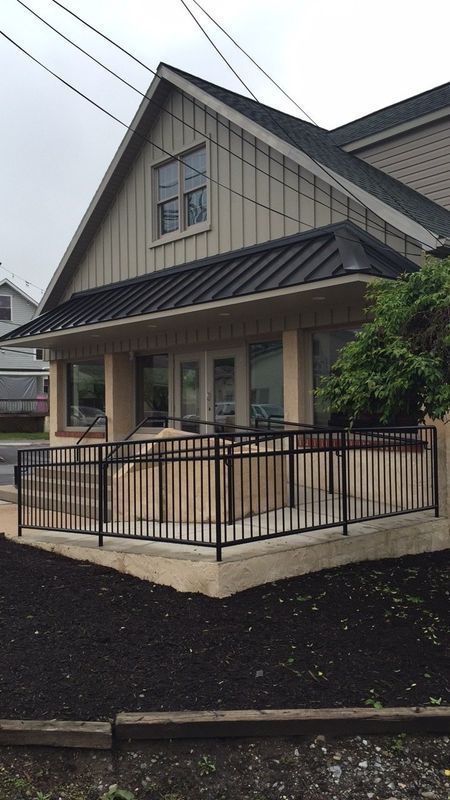 Exterior of a tan building with a black roof, front porch with black railing.
