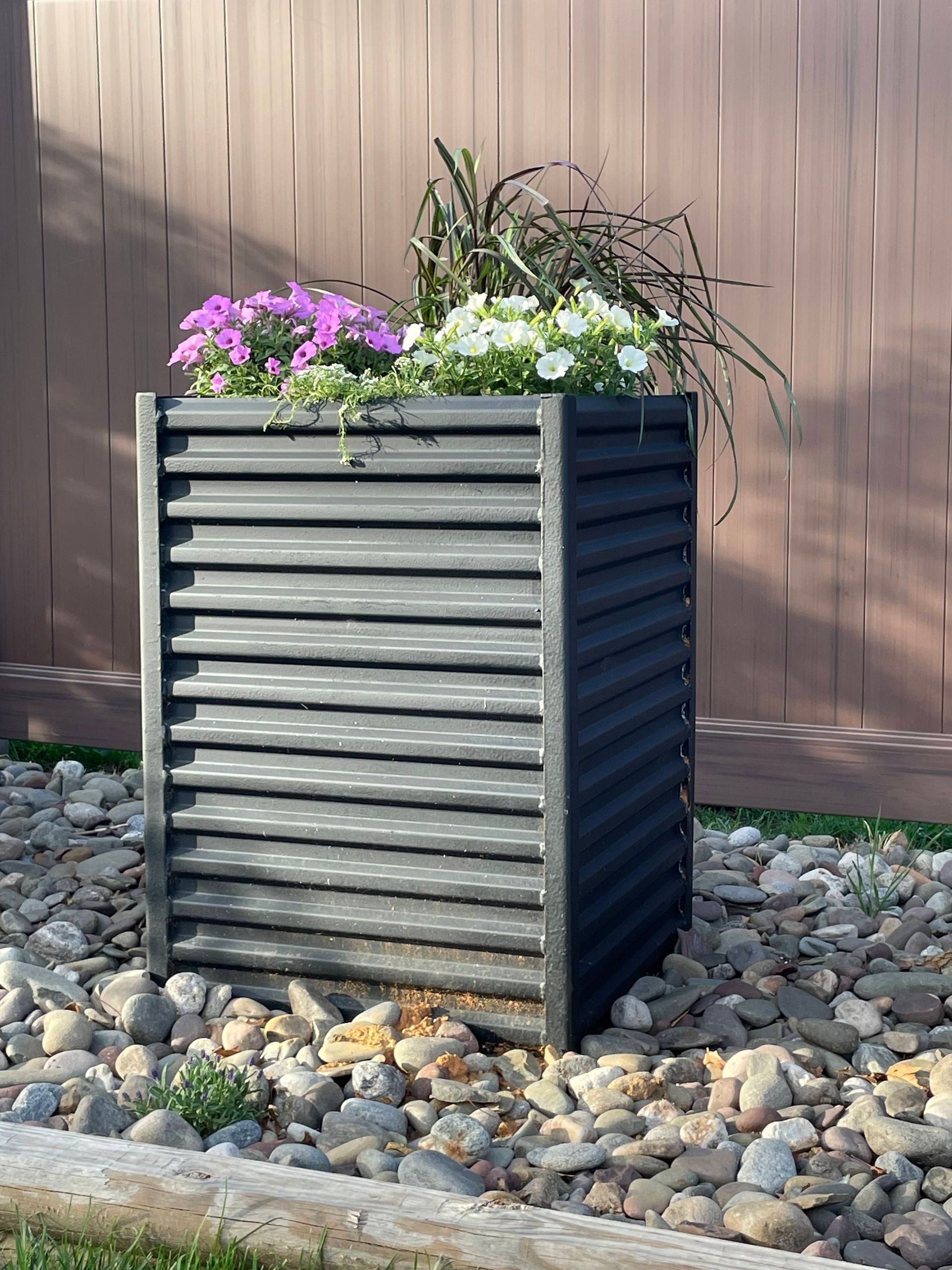 Black slatted planter box with colorful flowers, set on rocks against a brown fence.