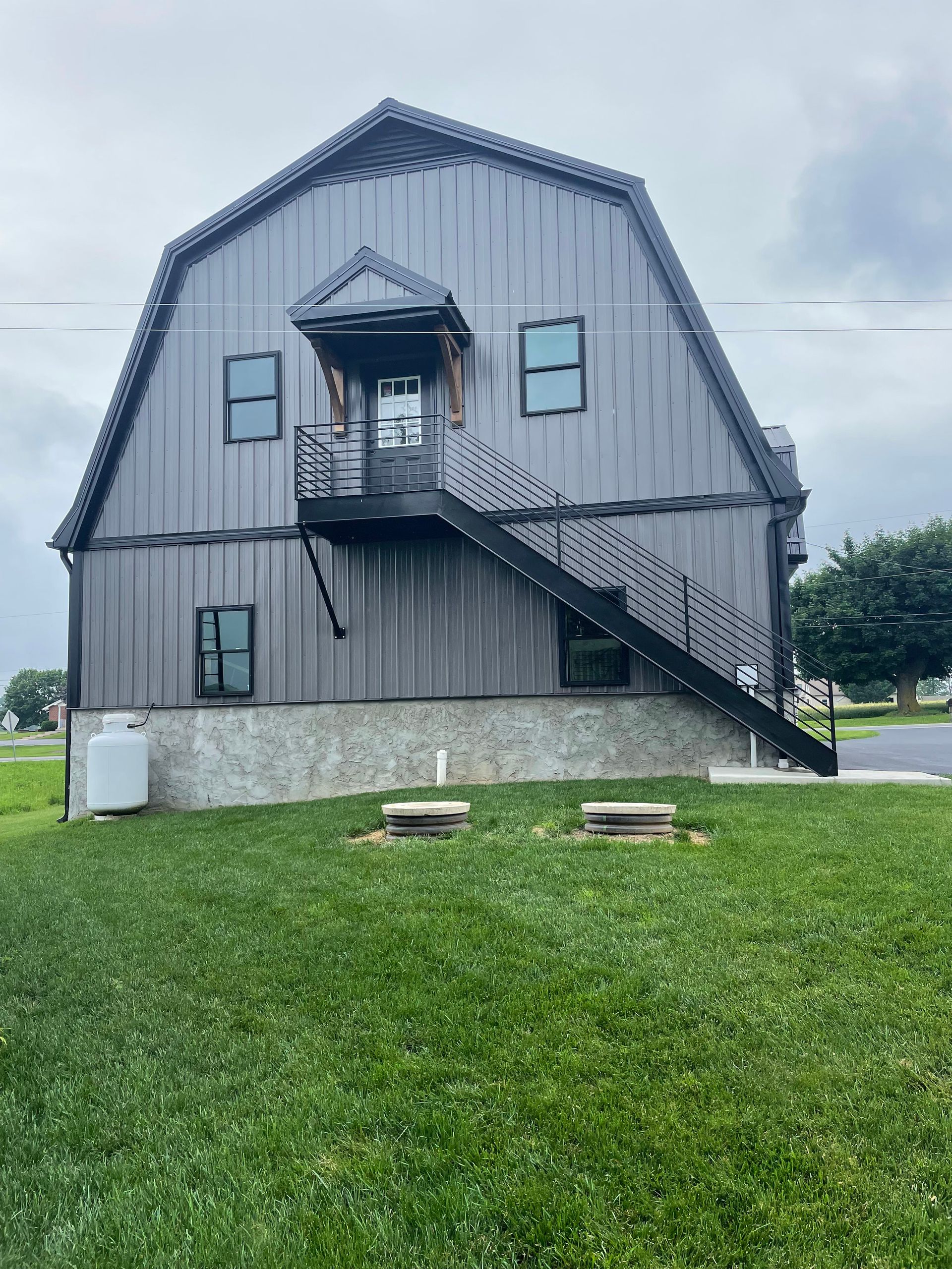 Gray metal barn-style building with exterior staircase and small porch. Green grass and overcast sky.