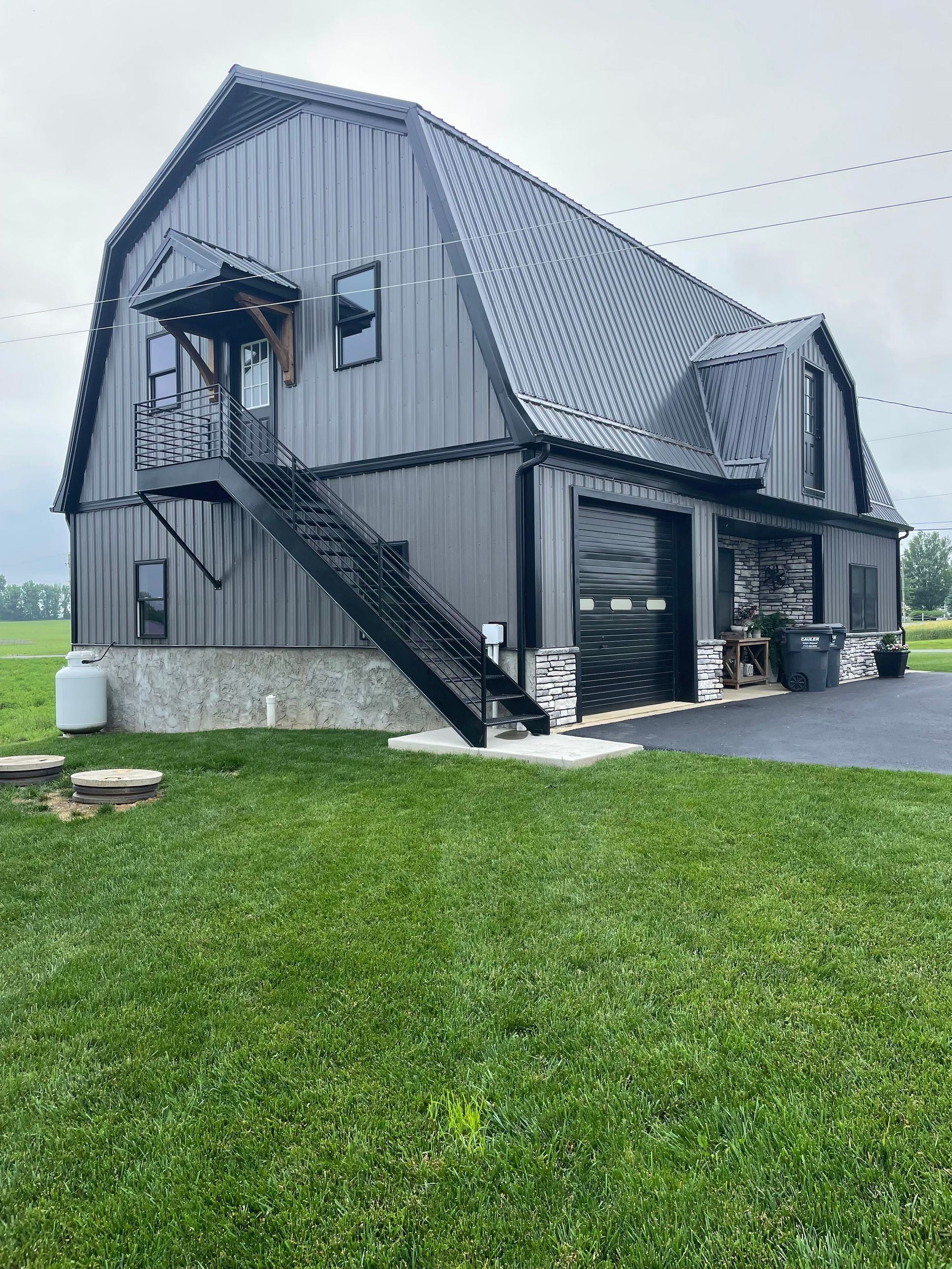 Modern, gray barn-style building with black doors, garage, and staircase, set on a green lawn.