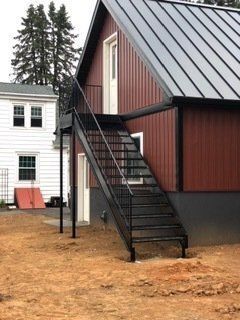 Black metal exterior stairs leading to a red building with a metal roof. Brown ground and a white building in the background.