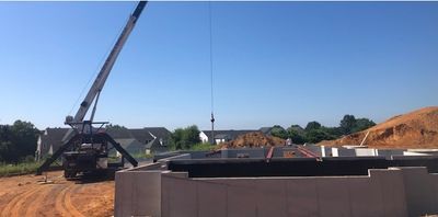 A crane lifts a large object over a concrete foundation on a construction site; blue sky overhead.