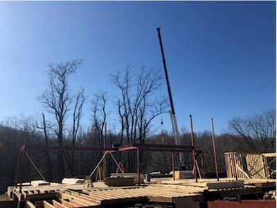 Construction site with a crane lifting a steel beam. Bare trees and blue sky in the background.