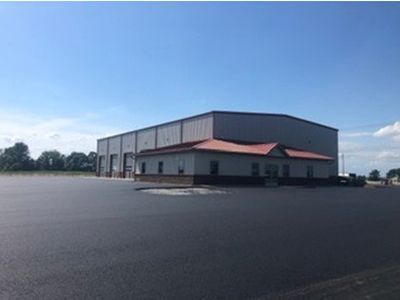 Large commercial building with a smaller red-roofed section attached. Paved parking area in front, blue sky overhead.