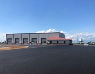 Large metal building with multiple garage doors, red-roofed side building, and asphalt parking area under blue sky.