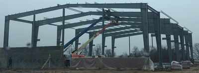 Construction of a steel-framed building under a cloudy sky, with workers and a lift in use.